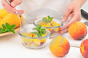 Chopped mangoes and peaches in a glass bowls with mint leaves close up on kitchen table, woman hands.