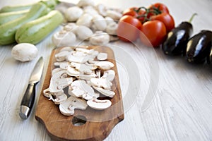 Chopped mushrooms on wooden chopping board and different vegetables over white wooden background, closeup. Side view.