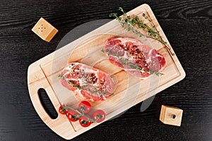 Chopped raw pork steaks with spices, tomatoes and thyme on a cutting kitchen board on a black wooden table. next to pepper shaker