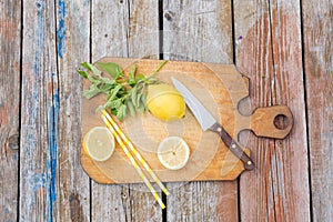 Chopping cutting board, sliced lemon, mint leaves, knife and straw on wooden table. Cocktail preparation. Summer. Top