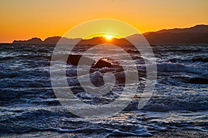 Choppy waves with boulders in water and golden sky with sun poking above mountains at sunset