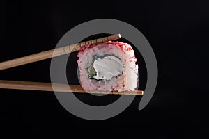 Chopsticks holding seaweed pink roll filled with avocado and fish over black background