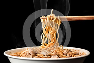 Chopsticks pick up tasty noodles with smoke on dark background. Ramen in white bowl.
