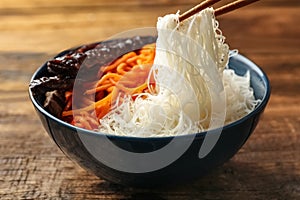 Chopsticks with tasty cooked rice noodles over bowl on wooden table, closeup