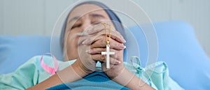 Christian cross in hands of cancer patient wearing headscarf during chemotherapy treatment lying on bed in hospital praying and