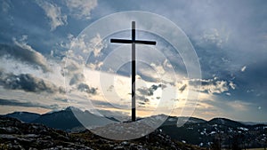 Christian cross on a rock in a mountain landscape, Dramatic clouds over the silhouette of the mountains in winter