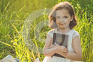 Christian girl holds bible in her hands. Reading the Holy Bible in a field during beautiful sunset. Concept for faith