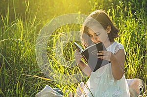 Christian girl holds bible in her hands. Reading the Holy Bible in a field during beautiful sunset. Concept for faith