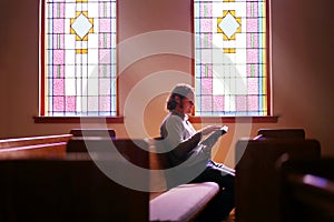 Christian Man Sitting Alone in Dark Empty Church Pew by Bright Stained Glass Window