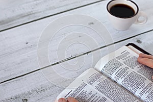 Christian woman holding and reading from open holy bible book placed on wooden table with a cup of coffee