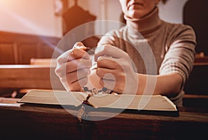 Christian woman praying in church. Hands crossed and Holy Bible on wooden desk.