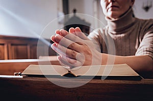 Christian woman praying in church. Hands crossed and Holy Bible on wooden desk.