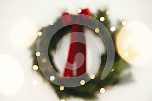 Christmas abstract background. Blurred stylish christmas wreath with red bow and golden lights on white wall. Merry Christmas and