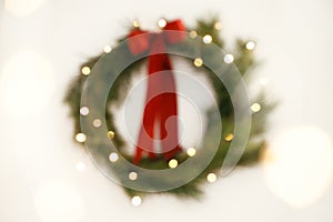 Christmas abstract background. Blurred stylish christmas wreath with red bow and golden lights on white wall. Merry Christmas!