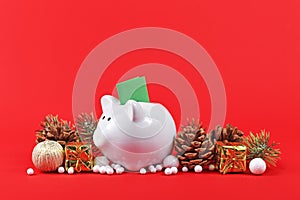 Christmas arrangement with piggy bank with green coupon surrounded by seasonal ornaments on red background