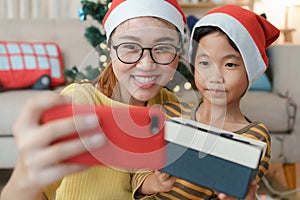 Christmas. Asian family with a mother and daughter giving gift box via video call together with smartphone in the living room at