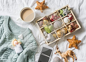 Christmas background. Box of vintage christmas decorations, coffee with milk, phone and blue knitted sweater on the bed, view from