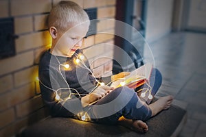 Christmas background. The boy is sitting near the fireplace with a book. Child with yellow lights. Waiting for the new year.