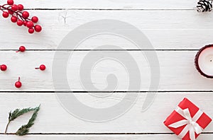 Christmas background with red gift box, holly berry, fir leaves, pine cone and candle on white wooden background.