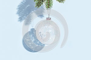 Christmas ball hanging on pine branch on blue colored wall. White glass transparent round New Year toy