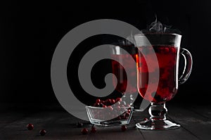 Christmas bright red mulled wine with berries, steam, ripe cowberry, blueberry in two glass wine glasses on black wood table.