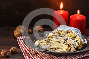 Christmas cake, german christstollen with fruits, raisins and marzipan in front of two red candles and nuts on rustic dark wood