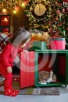 A Christmas card with a baby and a rabbit in a box under the Christmas tree. Christmas lights garlands and festive decor. A girl