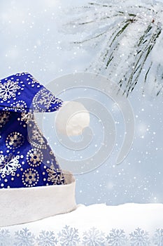 Blue decorated with snowflakes hat Santa Claus on snow on background of snow-covered pine branch and row of snowflakes.