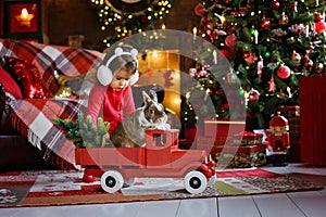 A Christmas card with a child and a rabbit in a toy car under a Christmas tree in red pajamas. Christmas lights garlands and