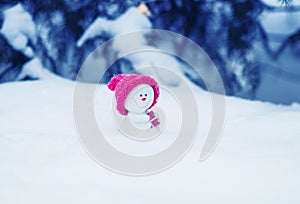 Christmas card with a funny toy snowman in a bright pink cap sitting in a snowdrift with a gift under the tree