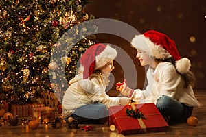 Christmas Child Girl opening Box and giving Gift Toy to Sister. Happy Children in Santa Hat next to Xmas Tree. Fairy Magic