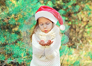 Christmas child little girl in santa red hat blowing snow lying on her hands over tree