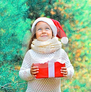 Christmas child little girl in santa red hat with gift box dreaming near tree