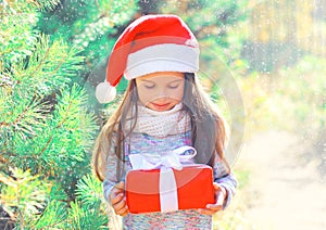 Christmas child little girl in santa red hat with gift box near branch tree