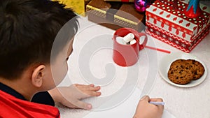 Christmas Child Write Letter to Santa Letter. Kid in Xmas Hat Writing Gift Wish List next to Christmas Tree over dark