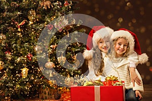 Christmas Children Sisters opening Present Box with Gift. Happy smiling little Girls in Santa Hat near to Christmas tree in dark