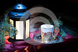 Christmas coffee cup and bright light glowing lantern, green fir tree branches.