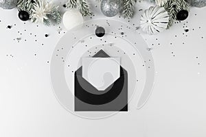 Christmas composition, black envelope, white and silver decorations, fir tree branches, silver stars confetti on white background.