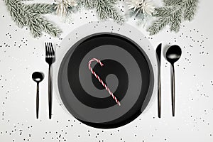 Christmas composition, black plate and cutlery, fir tree branch, candy cane, silver stars confetti on white background.