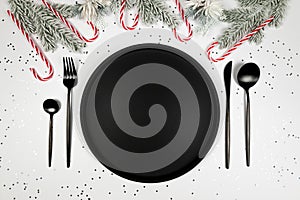 Christmas composition, black plate and cutlery, fir tree branch, candy cane, silver stars confetti on white background.