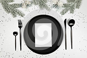 Christmas composition, black plate and cutlery, white card, fir tree branch, silver stars confetti on white background.