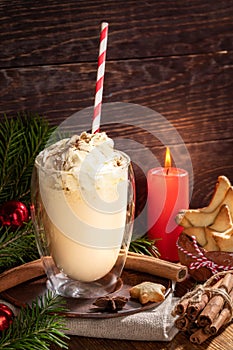 Christmas composition with eggnog with whipped cream in glass, candle, spices on wooden backdrop