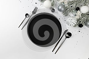 Christmas composition, white and silver decorations, black plate and cutlery, fir tree branches on white background.