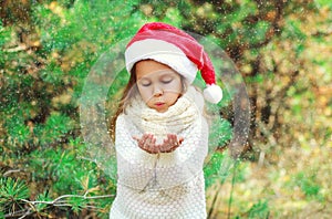 Christmas concept - little girl child in santa red hat blowing on snow in hands