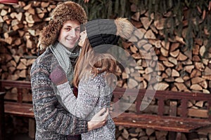 Christmas and couple concept - smiling man and woman in hats and scarf hugging over wooden country house and snow