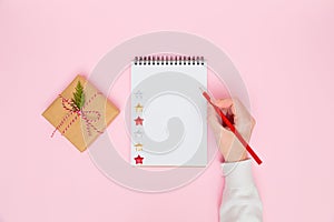 Christmas cozy background. Female's hand writing in blank notebook with craft gift box with red bow on pastel pink. New