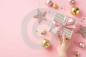 Christmas Day concept. First person top view photo of woman`s hand holding stylish present box with bow over gold pink baubles