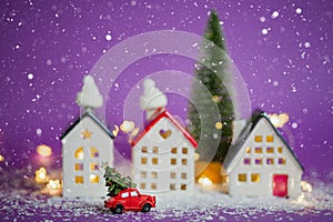 Christmas decor - red retro car on snow carries past houses with fairy lights in bokeh Christmas tree with gift boxes on roof. Toy
