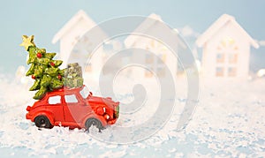 Christmas decor - red retro car on snow carries past white houses with lights garlands in bokeh Christmas tree with gift boxes on