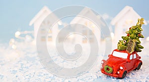 Christmas decor - red retro car on snow carries past white houses with lights garlands in bokeh Christmas tree with gift boxes on